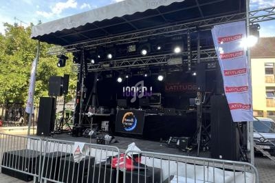 Scène extérieure équipée de matériel audio et éclairage, entourée de barrières, installée sur une place publique.