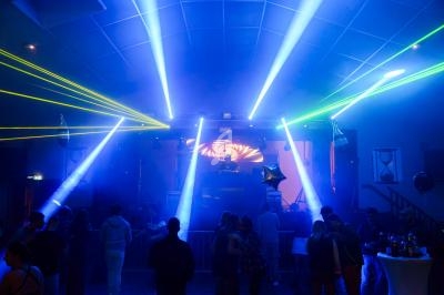 Salle de fête avec éclairages colorés, lasers et un DJ sur scène, entouré de personnes dansant et discutant.