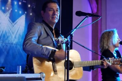 Un homme joue de la guitare sur scène avec une femme chantant au micro en arrière-plan.