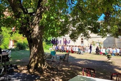 Un groupe de personnes assises à l'extérieur sous des arbres, près d'un bâtiment, avec du matériel audio visible au premier…