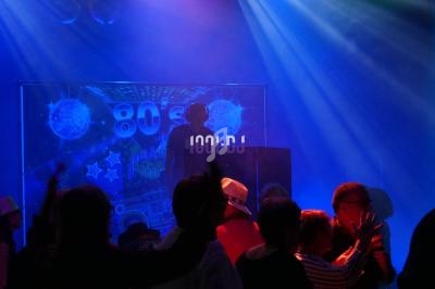 Personnes dansant sous des lumières colorées devant un DJ sur une scène décorée sur le thème des années 80.