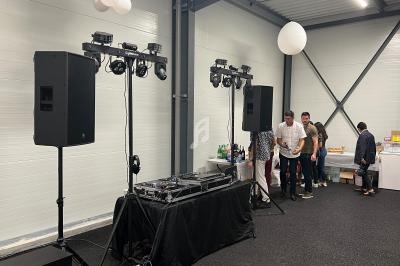 Deux grandes enceintes et une table de mixage installées dans une salle, avec des personnes rassemblées à l'arrière-plan.