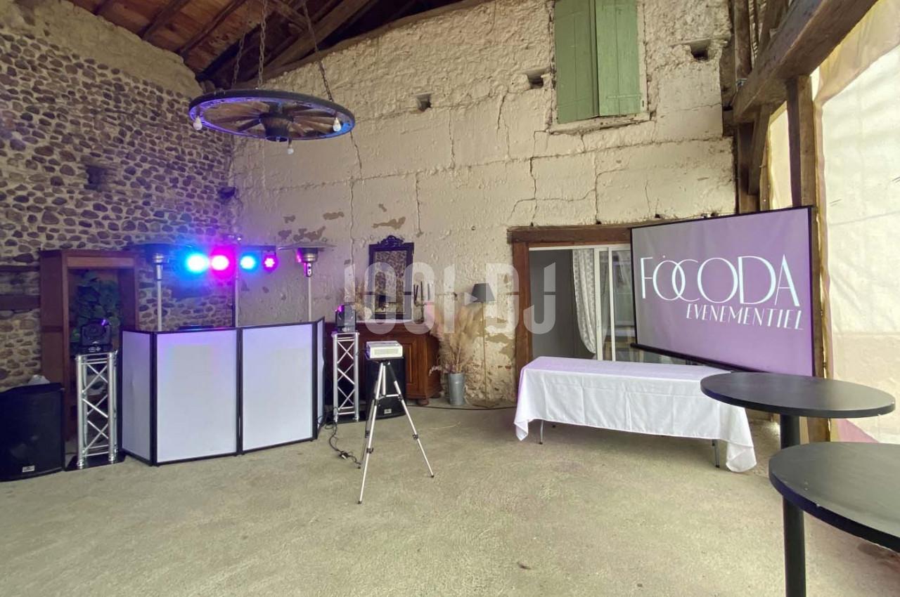 Salle rustique aménagée avec un espace DJ, éclairages colorés, tables hautes et écran affichant ’Foooda Événementiel’.