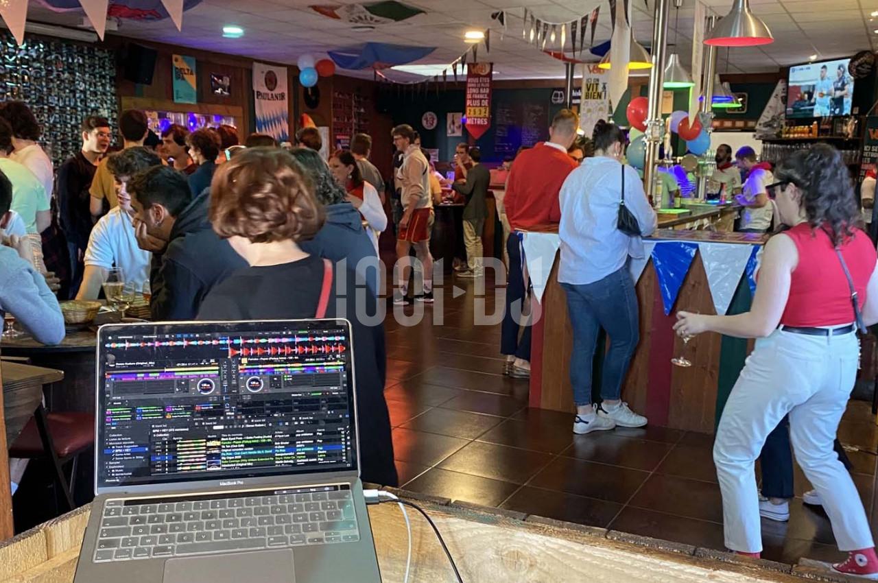 Des personnes discutent et boivent dans un bar décoré, avec un ordinateur affichant un logiciel de mixage au premier plan.