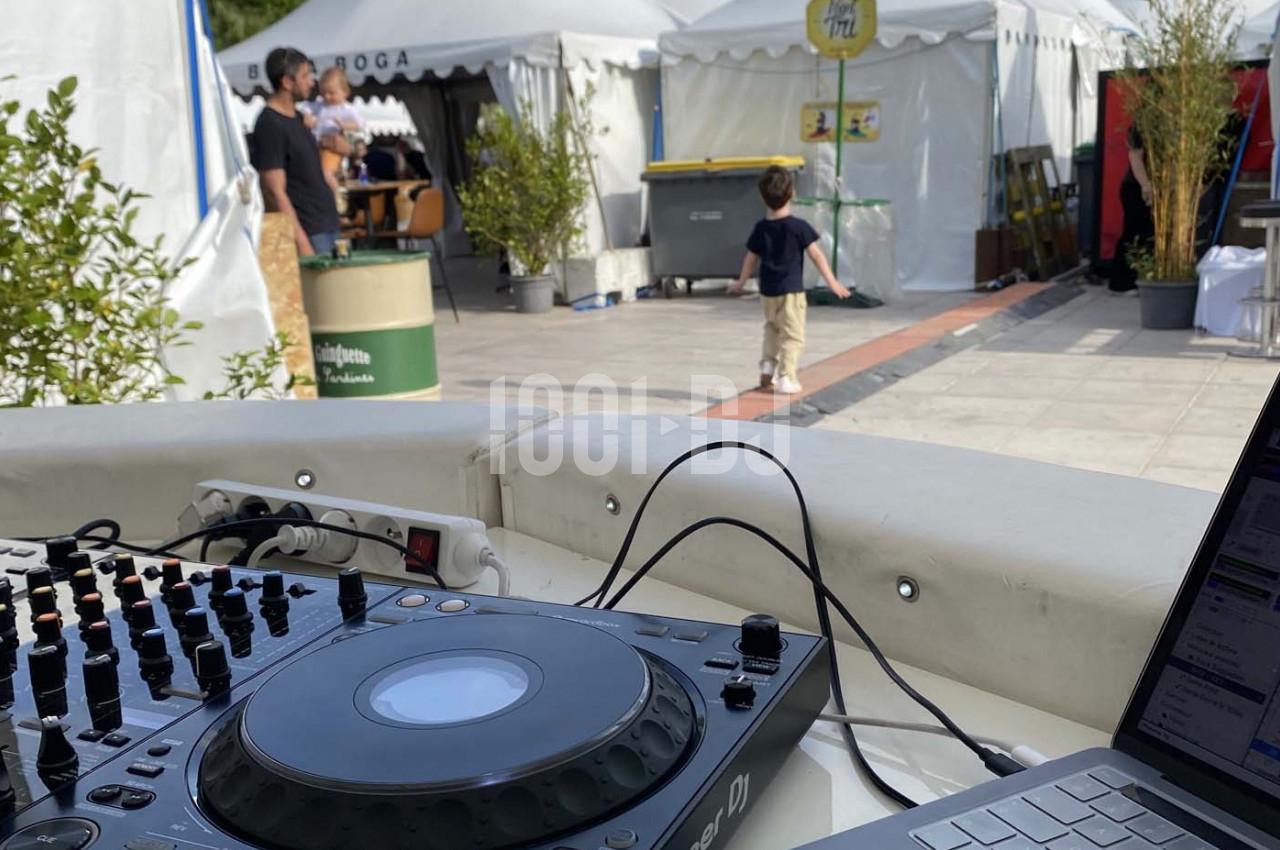 Vue d'une table de mixage et d'un ordinateur portable en extérieur, avec des stands et des personnes en arrière-plan.