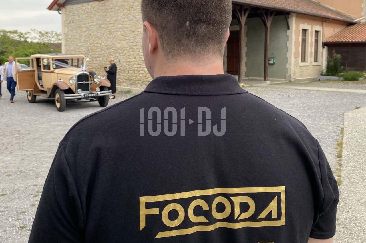 Un homme de dos portant un polo noir avec un logo doré, devant une voiture ancienne stationnée près d'un bâtiment en pierre.