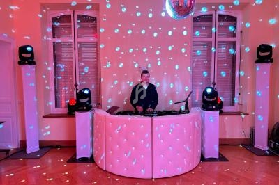 Un DJ mixe derrière une table blanche éclairée, entouré de lumières colorées et d'une boule à facettes.