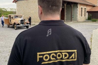 Un homme de dos portant un polo noir avec un logo doré, devant une voiture ancienne stationnée près d'un bâtiment en pierre.
