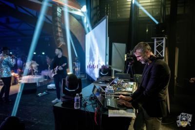 Un DJ mixe sur scène avec des musiciens en arrière-plan dans une salle éclairée par des projecteurs colorés.