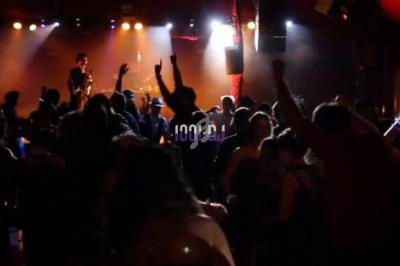 Foule dansante dans une salle sombre éclairée par des lumières colorées, avec un musicien sur scène en arrière-plan.