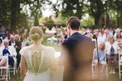 Un couple de dos, debout devant un micro, face à une assemblée en plein air lors d'une cérémonie.