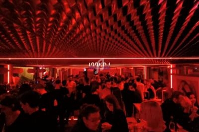 Salle de fête animée avec éclairage rouge, plafond texturé et personnes discutant autour de tables hautes.