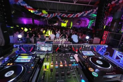 Vue depuis une table de mixage avec platines et ordinateurs, face à une foule dans une salle décorée de guirlandes colorées.