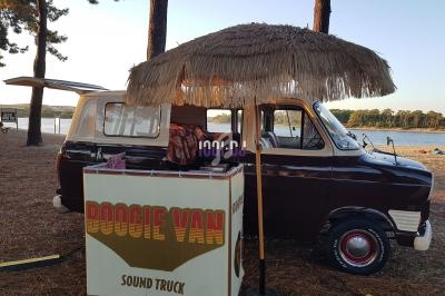 Un van vintage aménagé avec un comptoir et un parasol en paille, stationné près d'un lac entouré de pins.