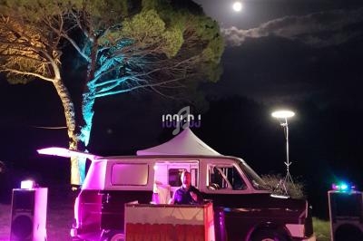 Un van aménagé en cabine de DJ avec éclairages colorés, installé en extérieur sous un arbre et une pleine lune.