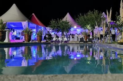 Vue nocturne d'une réception en plein air avec tentes éclairées, bassin au premier plan et invités en arrière-plan.