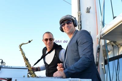 Deux musiciens jouent sur un bateau, l'un au saxophone et l'autre aux platines avec un casque audio.