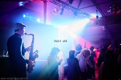 Un saxophoniste joue sur scène dans une ambiance festive avec des lumières colorées et des danseurs en arrière-plan.