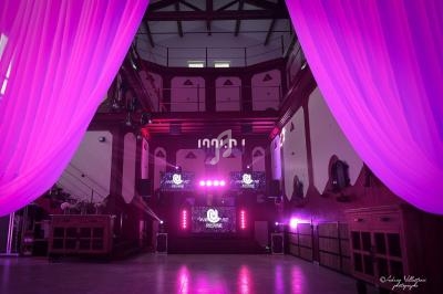 Salle de réception décorée de rideaux roses, éclairage violet et écrans lumineux sur une scène centrale.