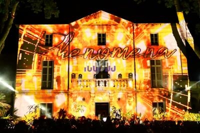 Façade d'un bâtiment illuminée par une projection lumineuse colorée avec le mot ’Phenomena’ visible.
