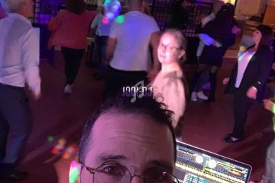 Un DJ anime une soirée dansante dans une salle éclairée par des lumières colorées, avec des participants en arrière-plan.