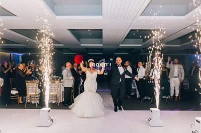 Un couple de mariés entre dans une salle de réception sous des étincelles, entouré d'invités applaudissant.