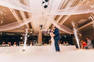 Un couple danse au centre d'une salle de réception décorée, entouré de jets d'étincelles et de tables ornées de fleurs.