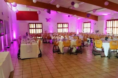 Salle de réception décorée avec des tables dressées, des chaises en bois et des lumières roses et violettes.