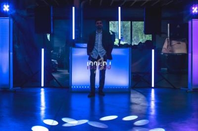 Un homme debout devant une table de DJ illuminée, entourée de lumières bleues et violettes dans un espace sombre.