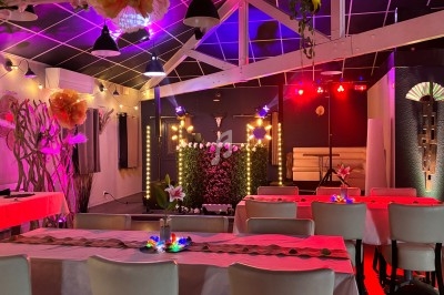 Salle décorée avec des tables dressées, éclairage coloré et scène ornée de guirlandes lumineuses et fleurs.