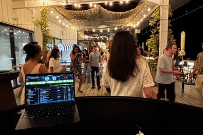 Vue depuis une table de mixage sur une terrasse illuminée, avec des personnes dansant et discutant en arrière-plan.