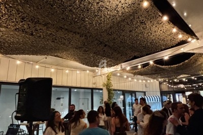 Personnes dansant et discutant sur une terrasse en soirée, éclairée par des guirlandes lumineuses et décorée d'un filet…