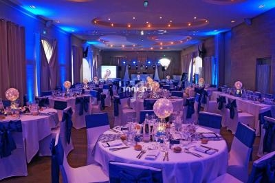 Salle de réception décorée avec des tables rondes, nappes blanches, chaises bleues et éclairage d'ambiance bleu.