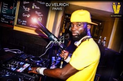 Un DJ portant une casquette jaune mixe sur des platines dans un bar avec des menus écrits à la craie en arrière-plan.