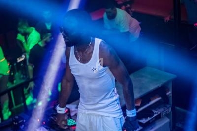 Un homme en débardeur blanc se tient près d'une table de mixage dans une ambiance de boîte de nuit.