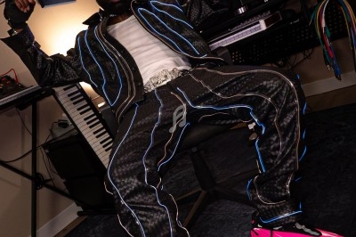 Un homme assis dans un studio d'enregistrement, portant une tenue noire lumineuse et des chaussures roses vives.