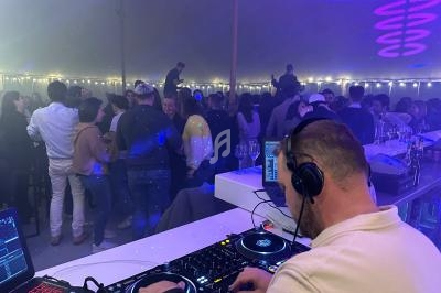 Un DJ mixe sur une table de mixage devant une foule dansante sous une tente éclairée par des guirlandes lumineuses.