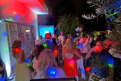 Groupe de personnes dansant en soirée en extérieur, éclairées par des lumières colorées, avec une table de mixage au premier…