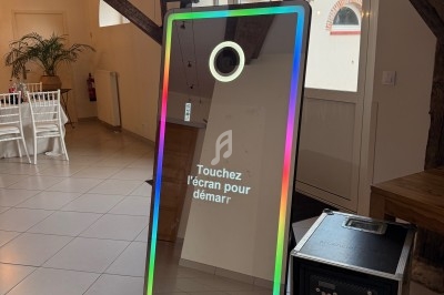 Borne photo interactive avec écran tactile et cadre lumineux, installée dans une salle avec poutres apparentes.