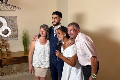 Un couple en tenue de mariage pose avec deux invités dans une pièce éclairée, décorée simplement.