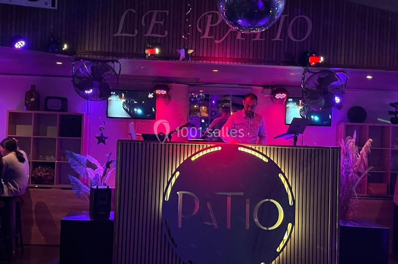 Un DJ mixe sur une scène éclairée par des lumières colorées dans un lieu nommé ’Le Patio’.
