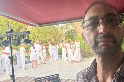 Un homme prend un selfie devant une terrasse où des personnes en tenue blanche discutent debout près de tables hautes.