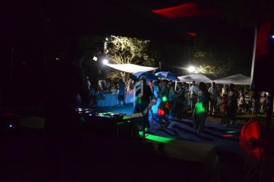 Personnes dansant sous des lumières colorées lors d'une fête en plein air, avec des stands éclairés en arrière-plan.