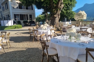 Tables rondes décorées de fleurs blanches installées en extérieur, près d'un bâtiment et d'un arbre, avec vue sur les…