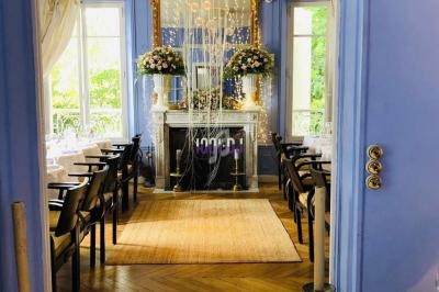 Salle de restaurant élégante avec murs bleus, cheminée décorée de fleurs et grandes fenêtres laissant entrer la lumière.