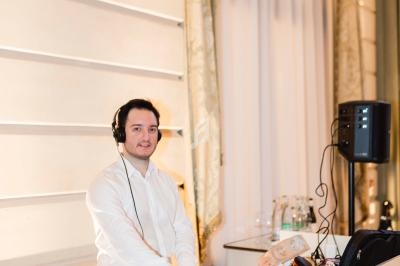 Un DJ en chemise blanche mixe de la musique derrière une table dans un intérieur élégant avec rideaux et haut-parleurs.