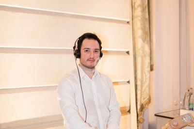 Un DJ en chemise blanche mixe de la musique derrière une table équipée de matériel audio dans une salle lumineuse.