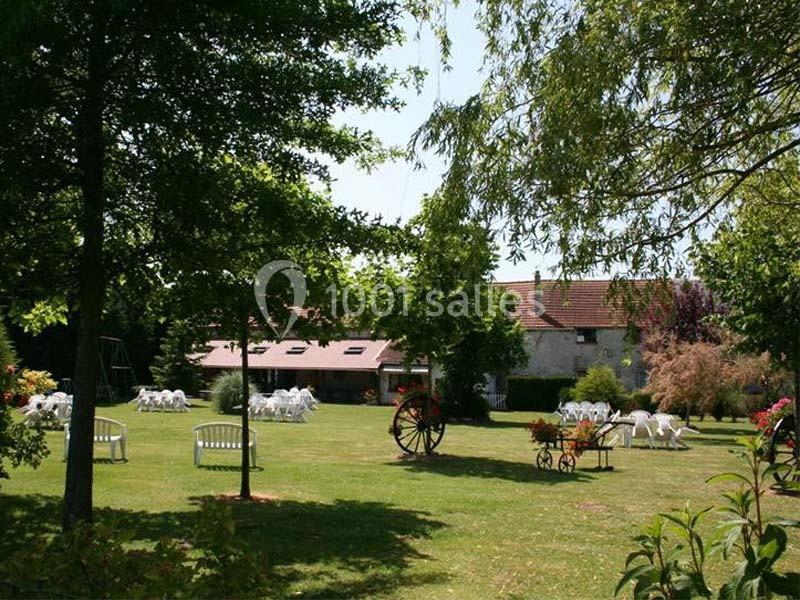 Jardin verdoyant avec des tables et des chaises blanches disposées près d'un bâtiment en pierre sous un ciel dégagé.