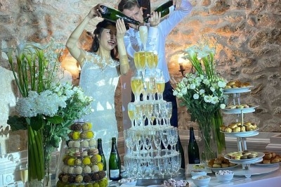 Un couple verse du champagne dans une pyramide de coupes sur une table décorée de fleurs et de desserts.
