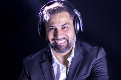 Homme souriant portant un casque audio et un costume sombre, éclairé par une lumière douce sur fond noir.